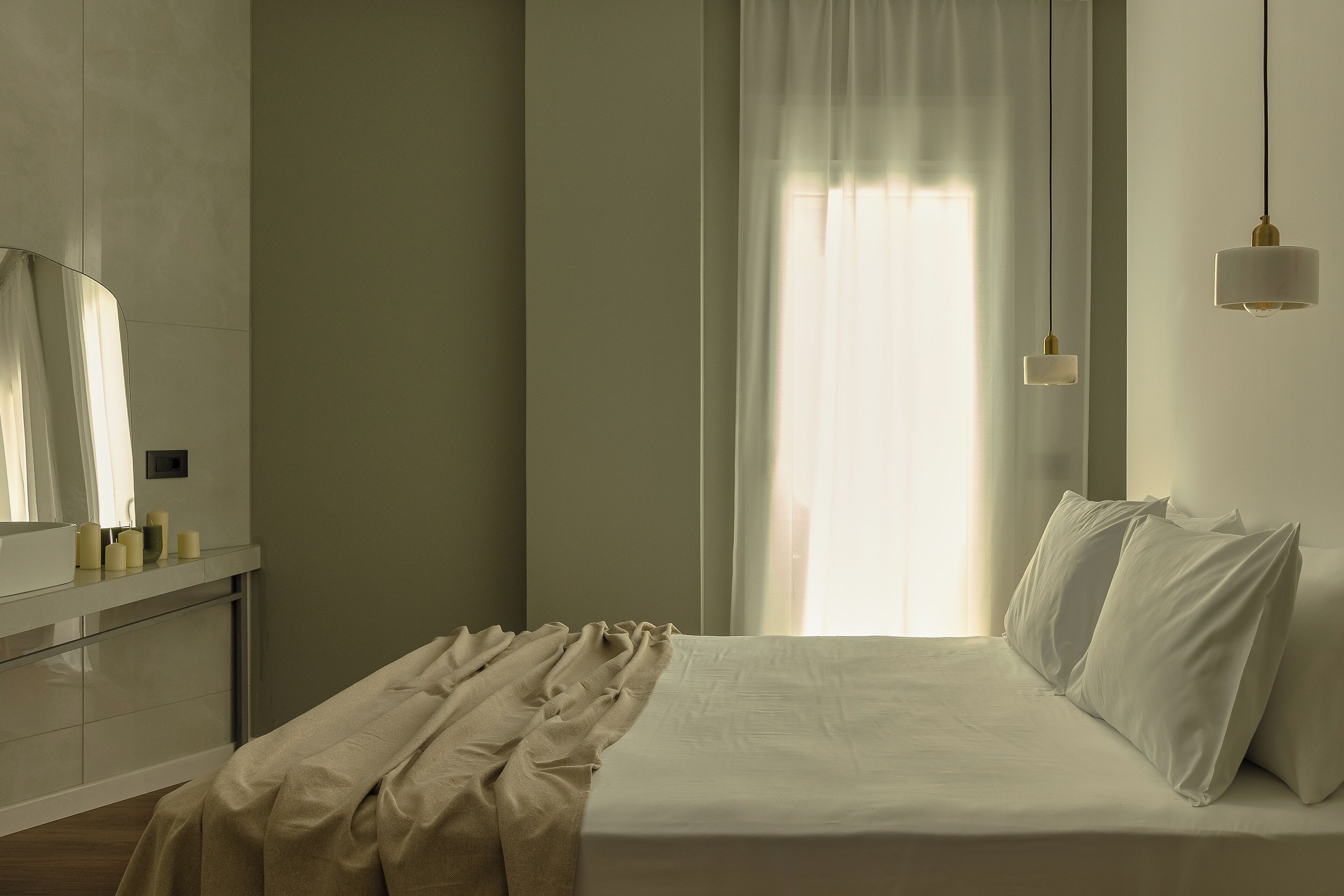 Un lit bien fait, avec du linge de lit blanc, une table de chevet dotée d’un miroir et de bougies, et une fenêtre ornée de rideaux légers laissant filtrer la lumière naturelle.