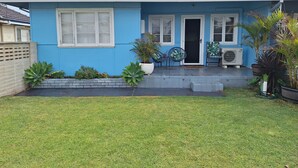 A single-story house with a blue exterior, a front porch with two chairs and a table, and a small garden area with various plants. - South Bunbury Beach Shack (South Bunbury)