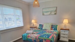 A bedroom with a bed, two bedside tables, a window with blinds, a hanging light fixture, and a wall-mounted painting. - South Bunbury Beach Shack (South Bunbury)