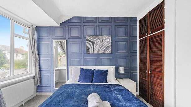 A bedroom with a bed, a window, a blue wall, a wooden door, and a marble artwork. - Cosy 2 Bed 5 Guests Garden Parking-wifi & Netflix (London)