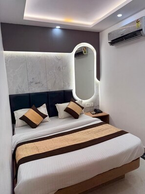 A modern hotel room with a large bed, a bedside table, a mirror, and an air conditioning unit. - Hotel Amba Deluxe (New Delhi)