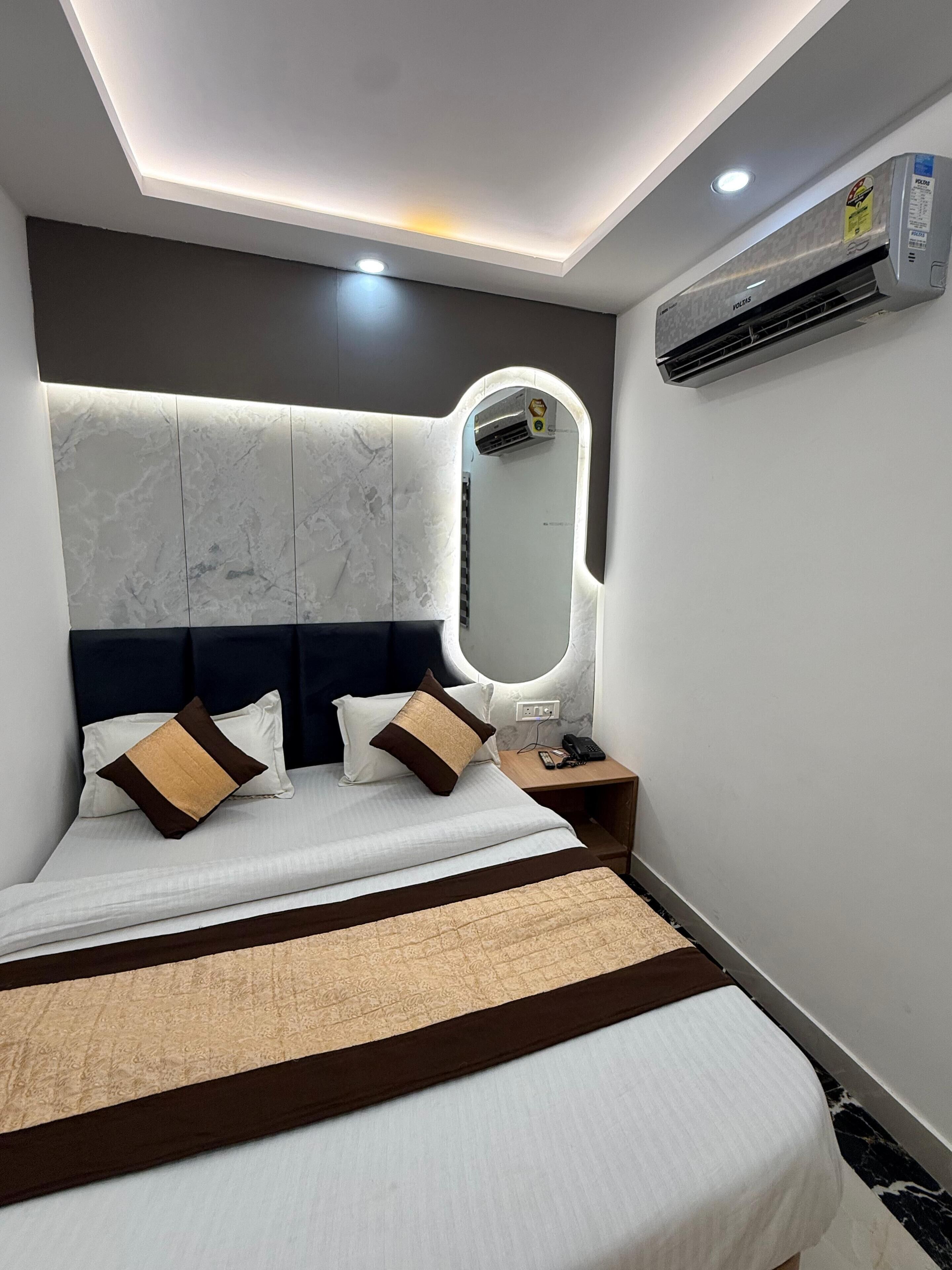 A modern hotel room with a bed, bedside table, mirror, and built-in air conditioning unit.