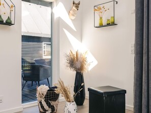 Een modern interieur met een glazen deur naar een terras, een zwarte leren poef, een rieten stoel en een vaas met gedroogde planten.