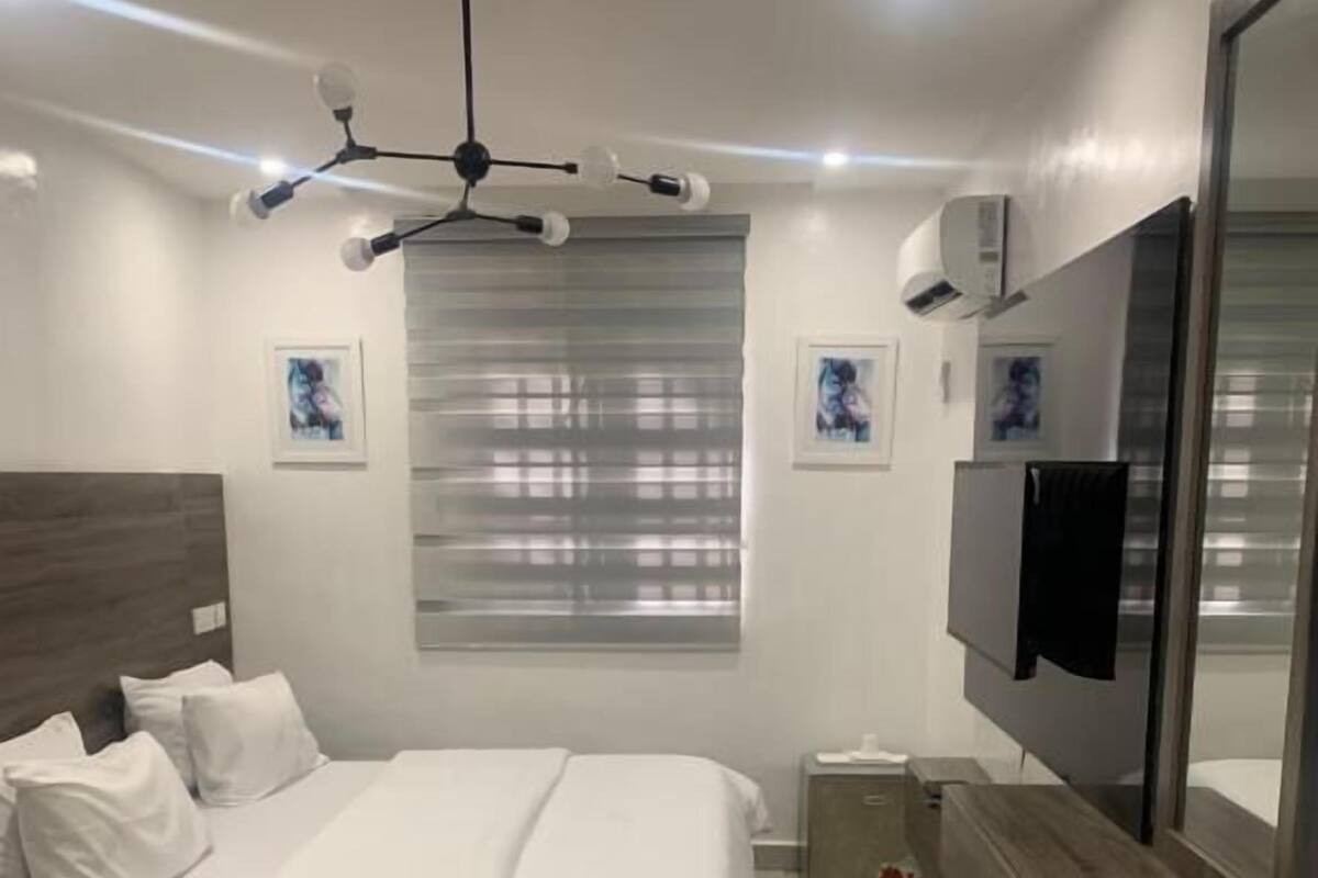 Une chambre moderne équipée d’un lit, d’une télévision, d’un ventilateur de plafond et d’une fenêtre avec des stores.