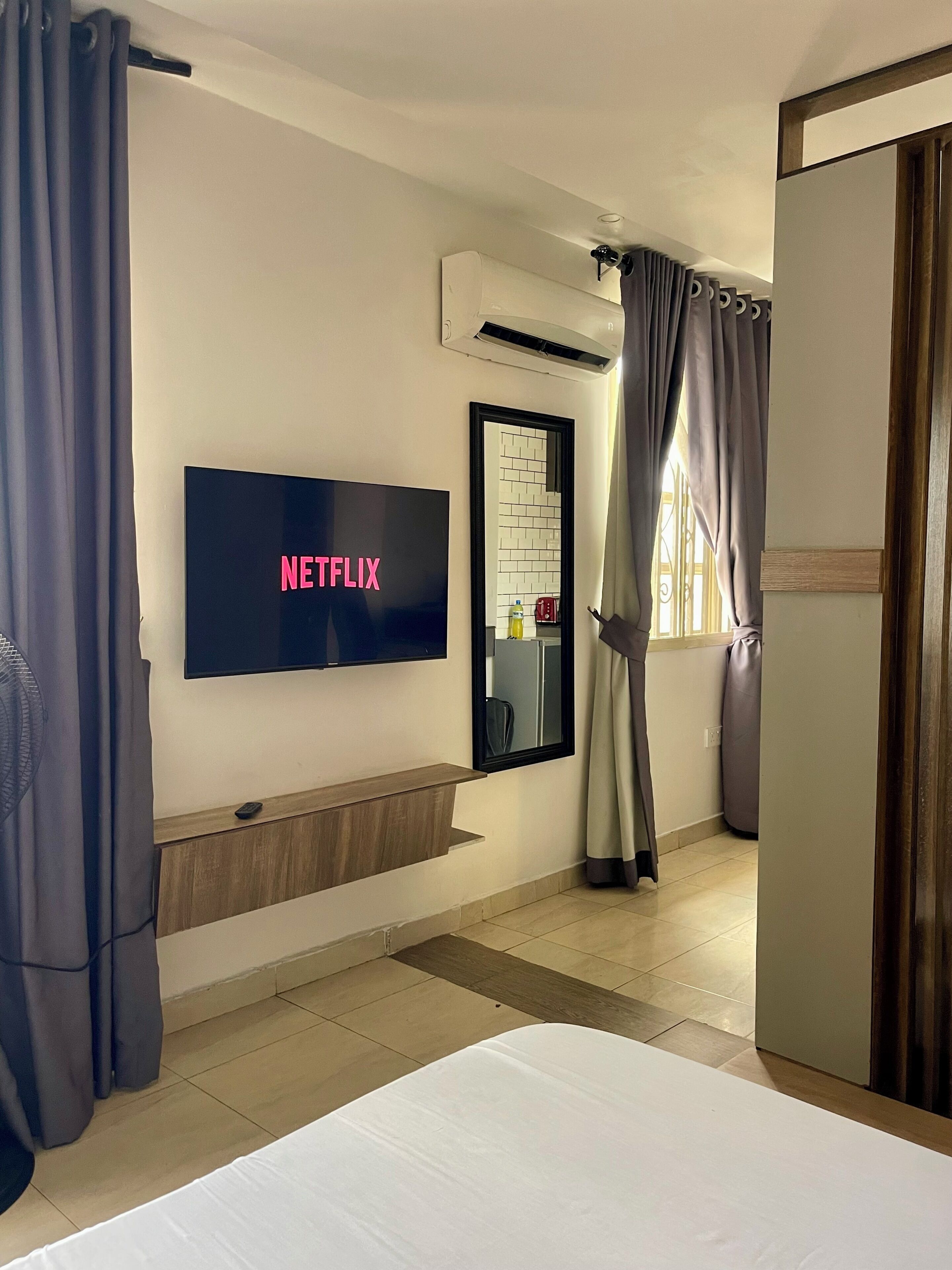 Quarto de hotel com uma cama, uma televisão na parede exibindo 'NETFLIX', um pequeno banco de madeira, um aparelho de ar condicionado e um espelho com painel de azulejos.