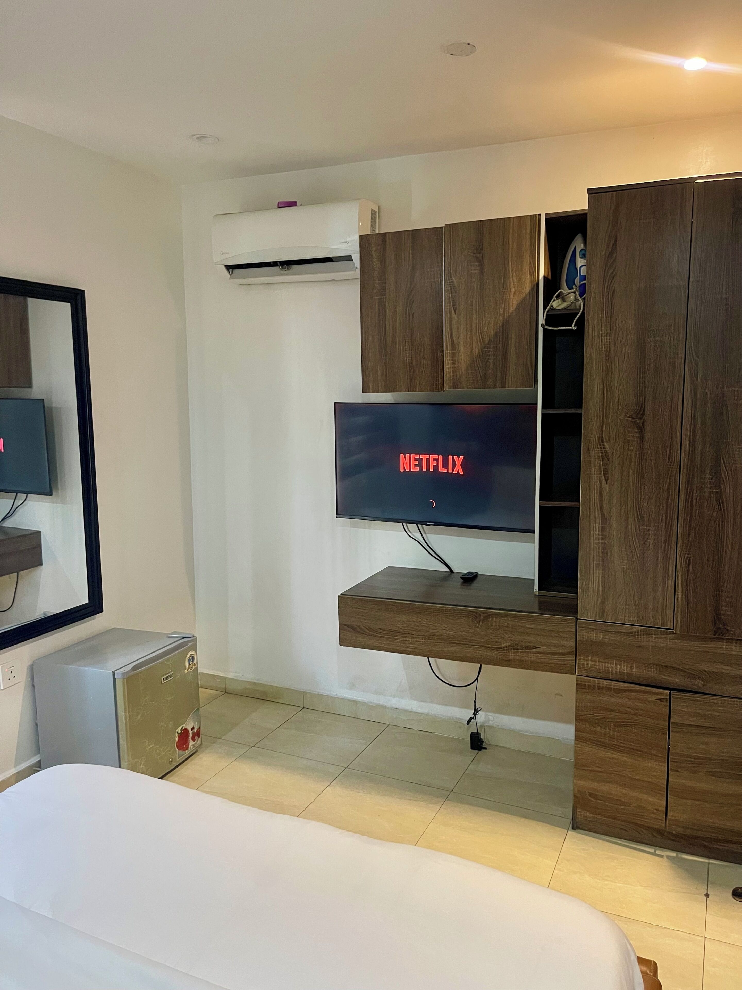 A modern hotel room with a flat-screen TV mounted on a wooden cabinet, a small bedside table, and a wall-mounted air conditioner.