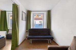 Ein kleines Zimmer mit einem Einzelbett, einem Fenster mit Vorhängen, einem Heizkörper und Blick auf ein Gebäude.