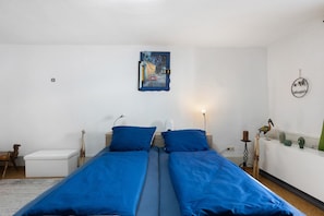 Ein Zimmer mit zwei Betten, einem Nachttisch, einer Lampe, einem kleinen Tisch mit einem Kaktus und einer Wanduhr.