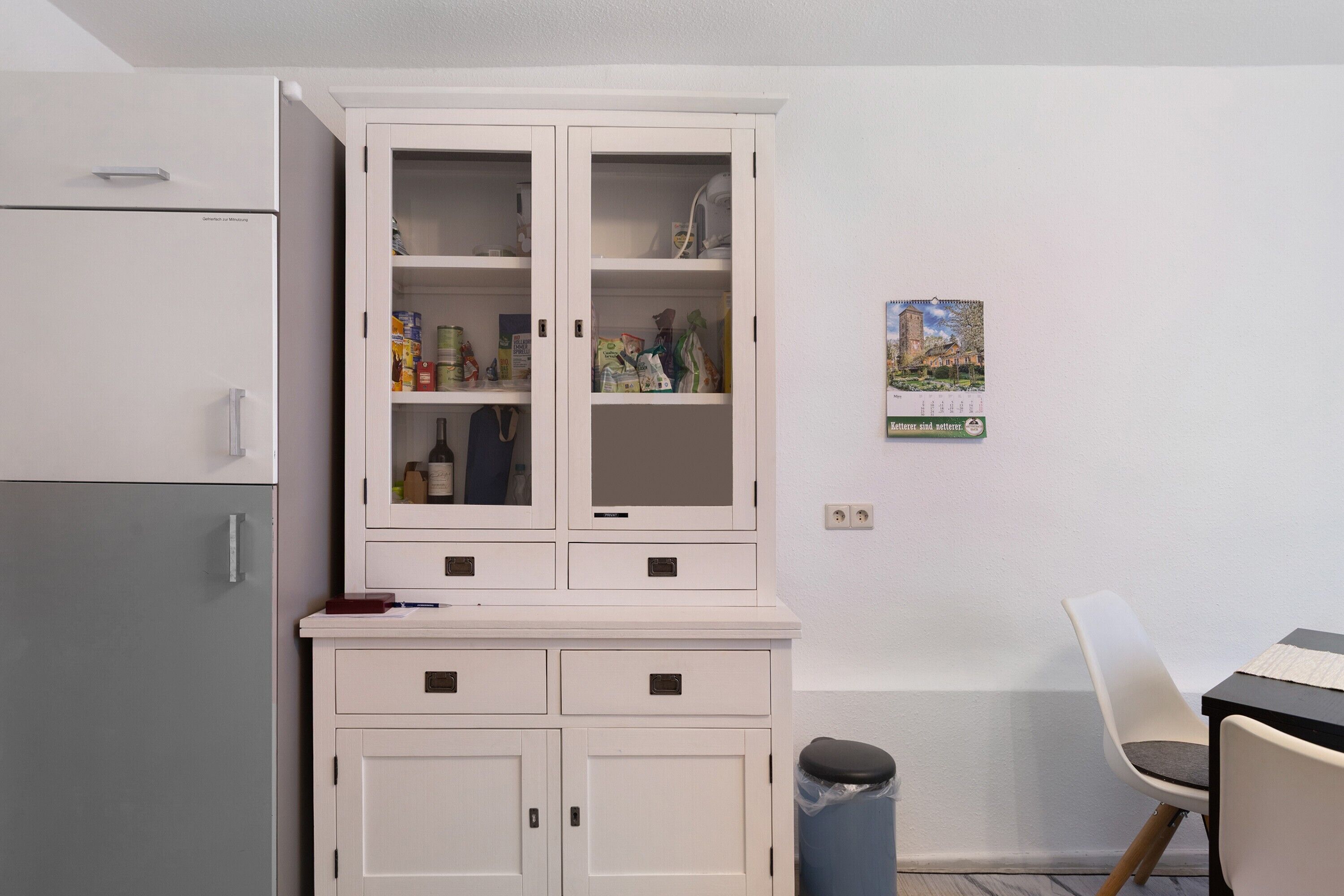 Een kitchenette met een koelkast, een witte kast met glazen deuren, een prullenbak en een stoel.