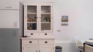 Een kitchenette met een koelkast, een witte kast met glazen deuren, een prullenbak en een stoel.