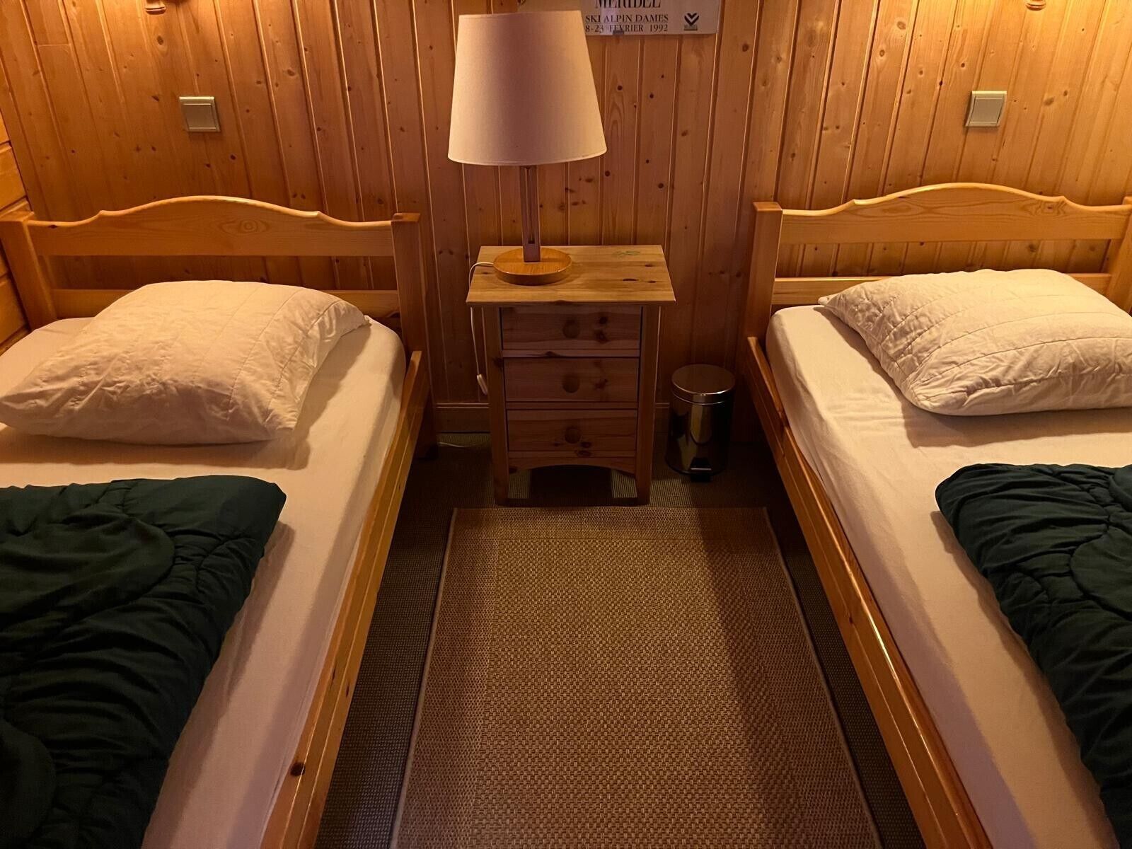 A room with two wooden beds, a nightstand with a lamp, a small drawer, and a trash bin.