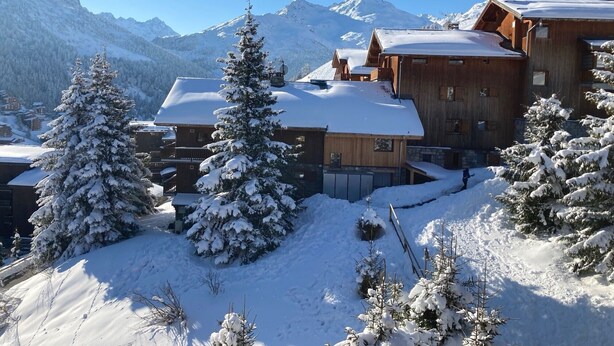 Un albergue de montaña cubierto de nieve con arquitectura de madera, rodeado de árboles y montañas cargadas de nieve.
