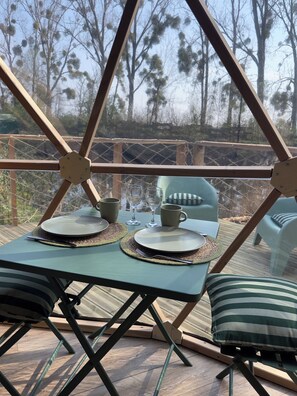 A dining area with a table set for two, surrounded by a wooden deck and a view of trees.