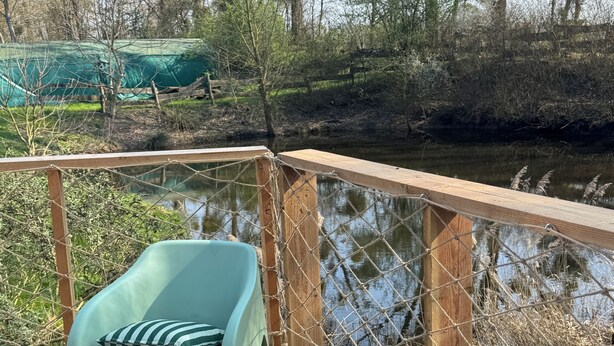 A wooden deck with two light blue plastic chairs and a cushion, overlooking a body of water and trees. - Nature Dome & Jacuzzi at Blue Grass Ranch (Bischoffsheim)