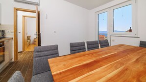 Ein modernes Zimmer mit einem großen Holztisch, grau gepolsterten Sesseln, einer Küchenzeile und großen Fenstern mit Blick.