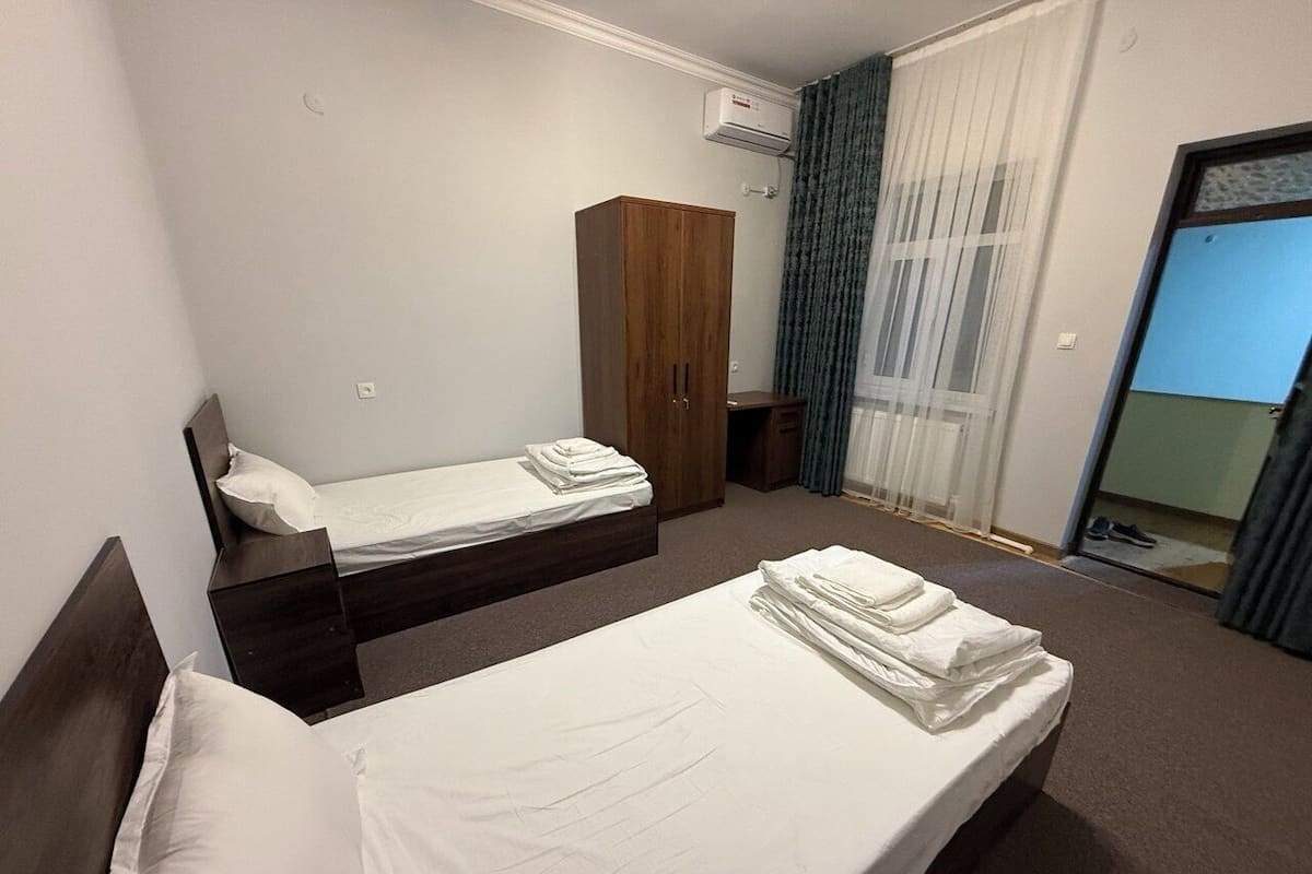 A hotel room with two beds, a wardrobe, an air conditioning unit, and a view of the outside through a window.