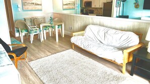Un salon comprenant un canapé, des fauteuils, une table basse, un tapis et un coin cuisine visible à travers une cloison en verre.