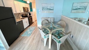 Une cuisine compacte avec des meubles blancs, un réfrigérateur noir et un four à micro-ondes. On y trouve également un coin repas avec une table en verre et des chaises blanches ornées de motifs verts en forme de feuilles.