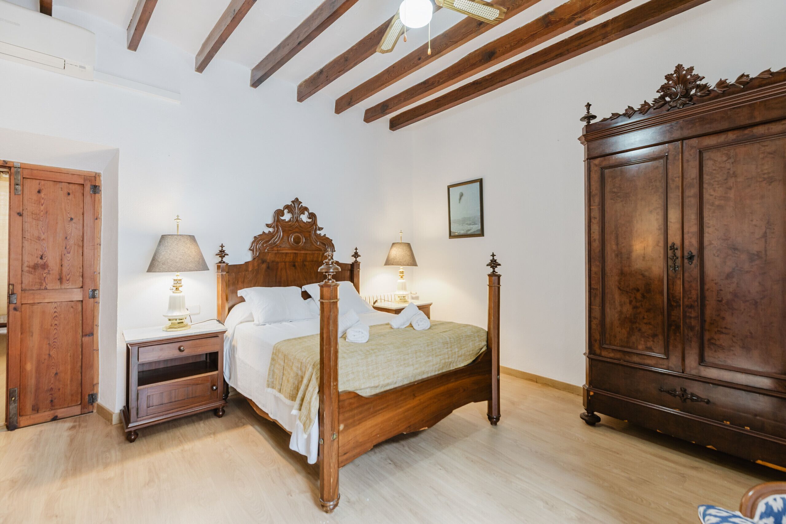 A bedroom with a wooden bed, two bedside tables with lamps, a wardrobe, and a ceiling with exposed beams.