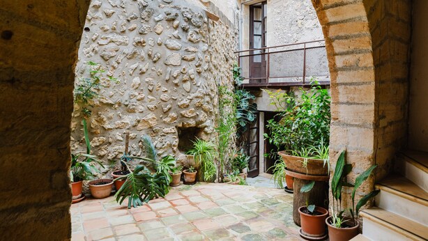 Une cour intérieure en forme d’arche, avec des murs en pierre, des plantes en pot et une porte en bois.