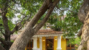 A courtyard with a yellow building, white columns, and a tiled roof. - amã Stays & Trails Chikoo Villa, Goa (Anjuna)