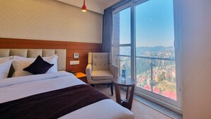 Habitación de hotel con cama, una silla, una mesita, una lámpara colgante roja y un ventanal con vistas a la ciudad.