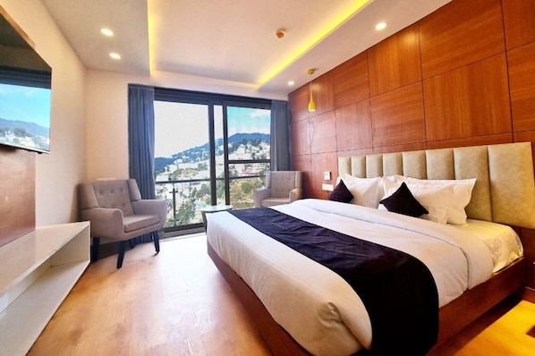 Quarto de hotel com uma cama grande, área de estar com duas cadeiras, televisão na parede e vista para a paisagem urbana através de uma janela ampla.