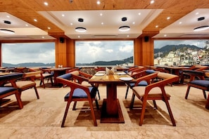 A restaurant with wooden tables and chairs, large windows, and a view of a mountainous landscape. - Mountain Valley View By Terraa (Shimla)