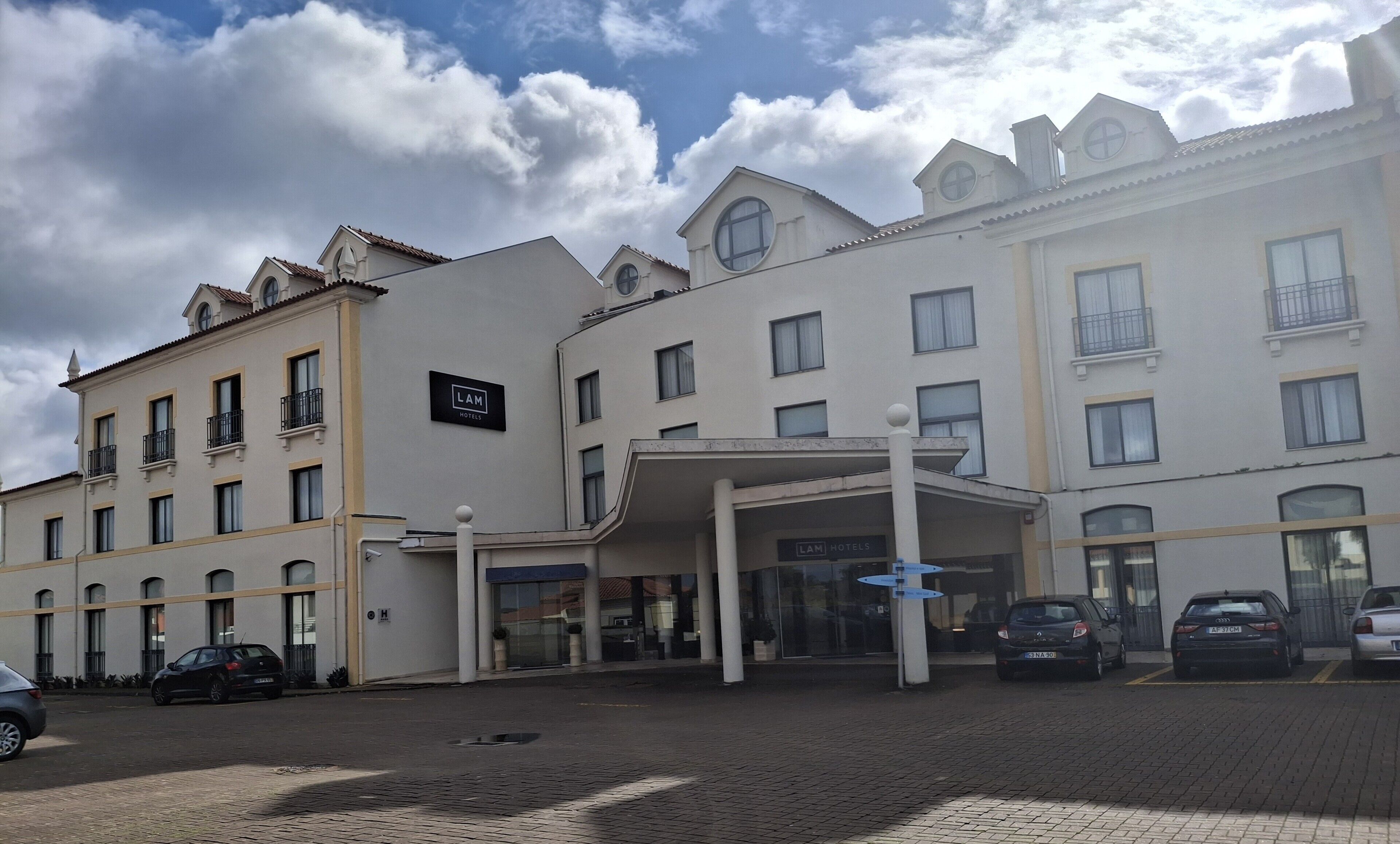 A hotel entrance with a covered area, multiple windows, and a parking lot with cars.