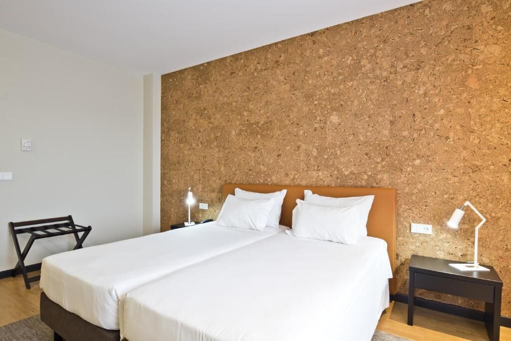 Habitación de hotel con una cama, dos mesitas de noche, una silla y un panel de corcho.
