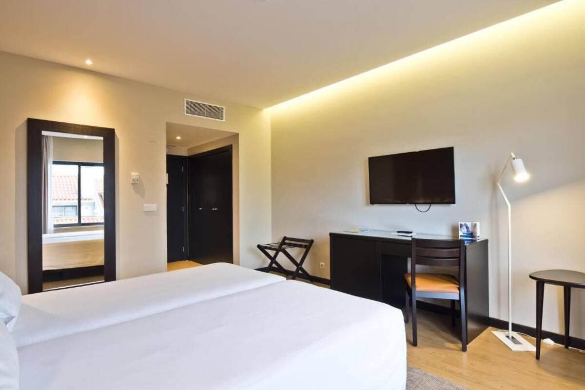 A hotel room with a bed, a desk, a chair, a TV, and a lamp.