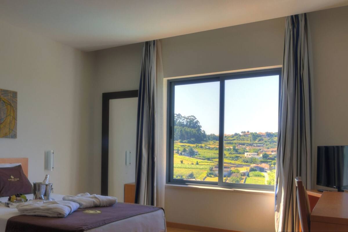 A hotel room with a large window, a bed with a headboard, a bedside table, a wall-mounted artwork, and a view of a landscape.