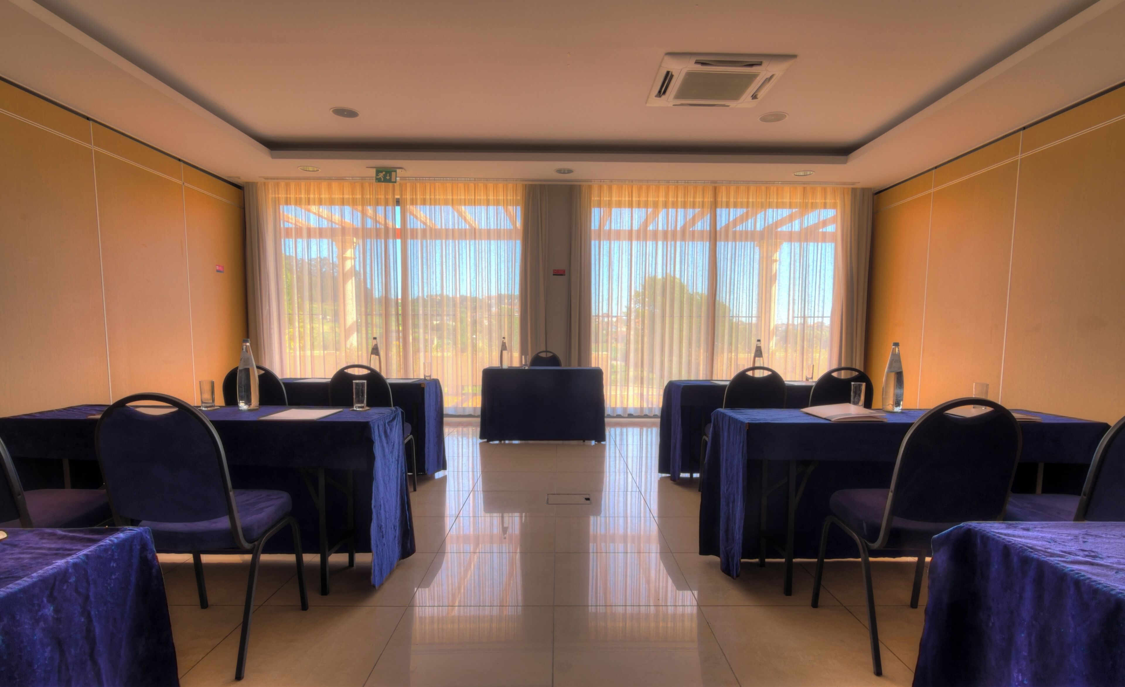 A conference room with tables and chairs, large windows with curtains, and a tiled floor.