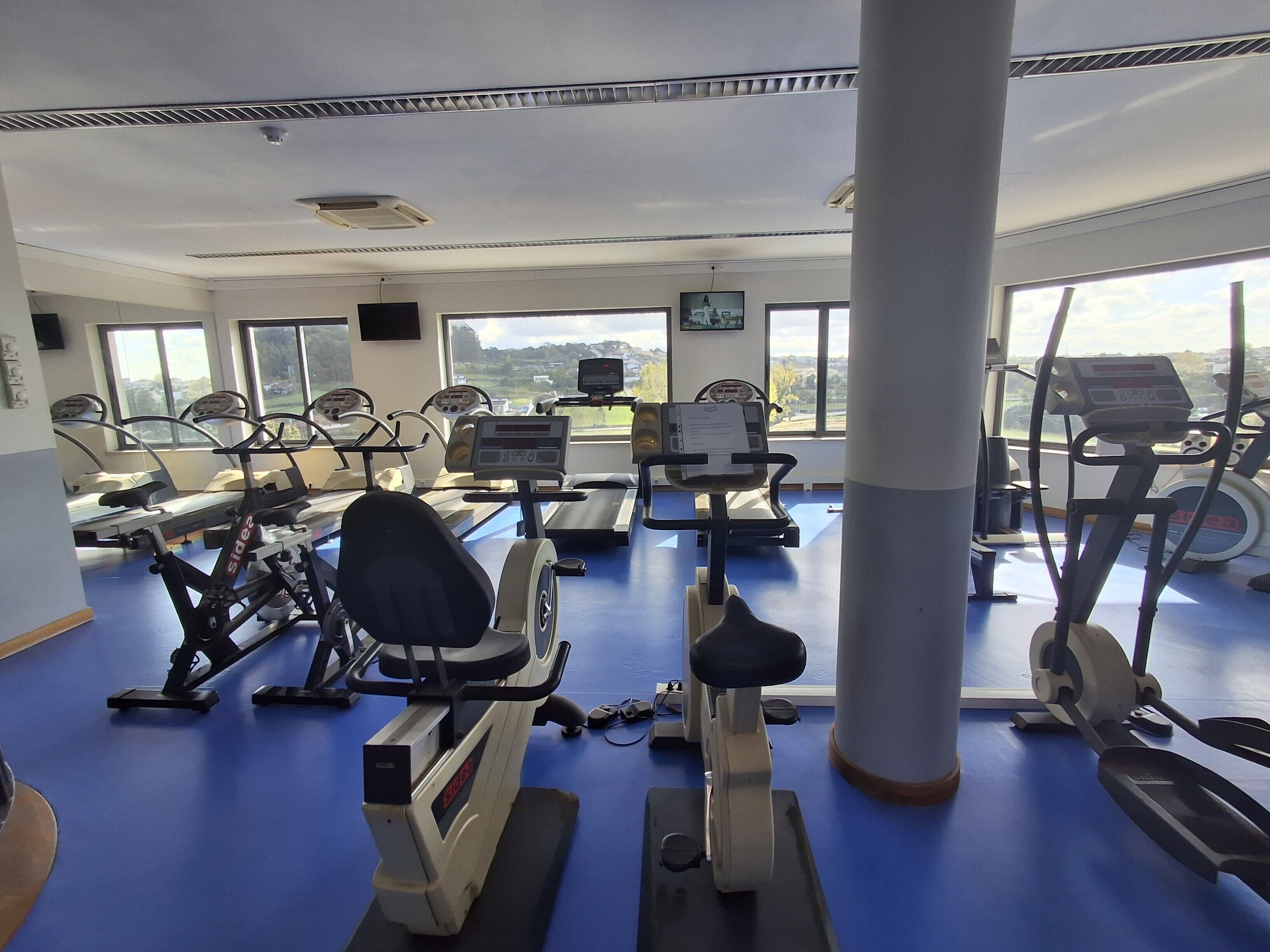 Uma academia com diversos equipamentos de ginástica, janelas amplas e vista para o exterior.