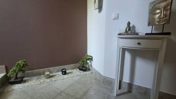 A room with a white console table, a Buddha statue, two potted plants, a small white bowl, and a wall-mounted light switch.
