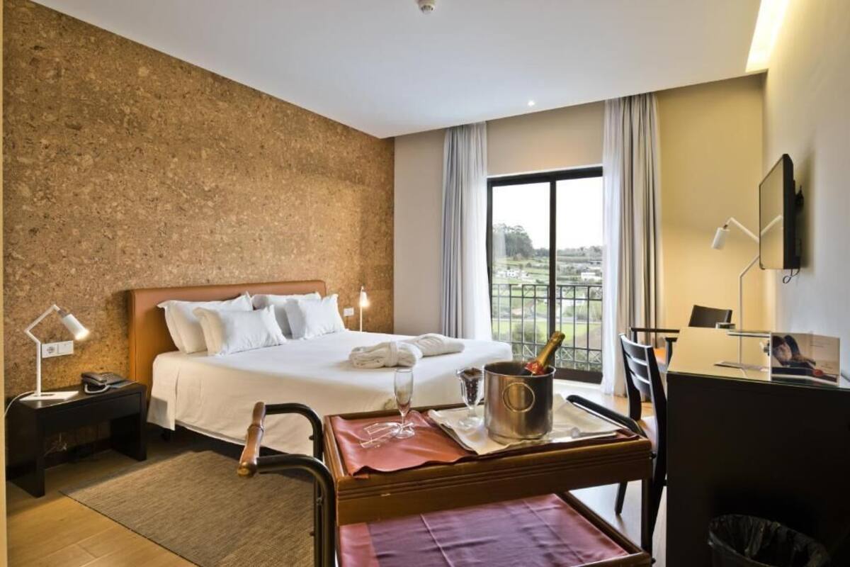 A hotel room with a bed, a desk, a chair, a trash bin, a nightstand, a lamp, a window, and a view of the outdoors.
