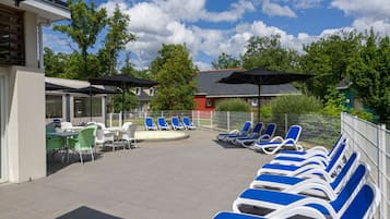 Ett område vid poolen med solstolar och bord, en överbyggd sittplats och en byggnad med ett sluttande tak.