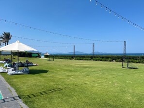 Ett grönområde utomhus med solstolar och parasoller, ett fotbollsmål och ljusslingor mot en klar himmel.