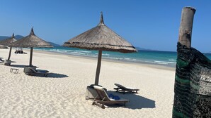 En strand med parasoller av halmtak, solstolar och fri utsikt över havet.