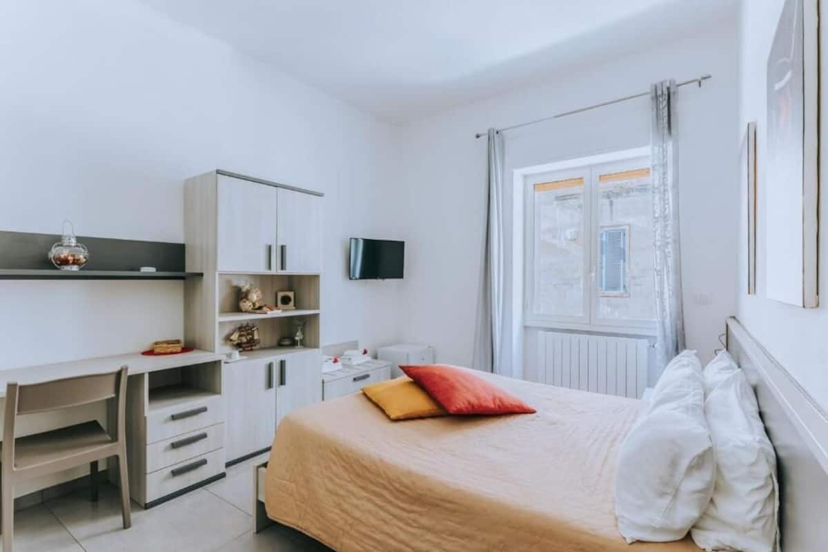 Een slaapkamer met een bed, een bureau, een televisie en een raam.