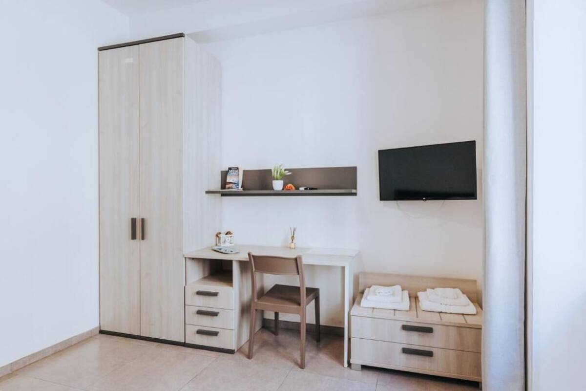 Een kamer met een garderobe, een bureau, een stoel, een aan de muur bevestigde televisie en een nachtkastje met een lamp.
