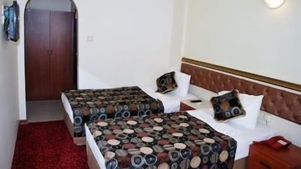 Habitación de hotel con dos camas, alfombra roja, puerta de madera y lámpara fijada a la pared.