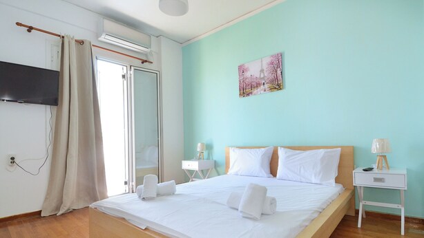 A hotel room with a bed, a flat-screen TV, a painting, a nightstand, and a window with curtains. - A cute & comfortable 2 bedroom apartment (Piraeus)
