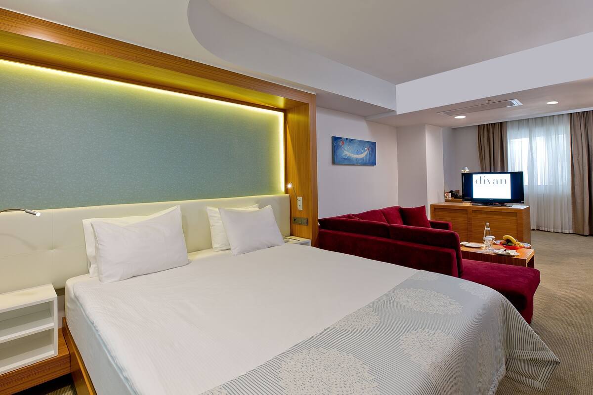A hotel room with a large bed, a desk with a TV, a red chair, and a painting on the wall.