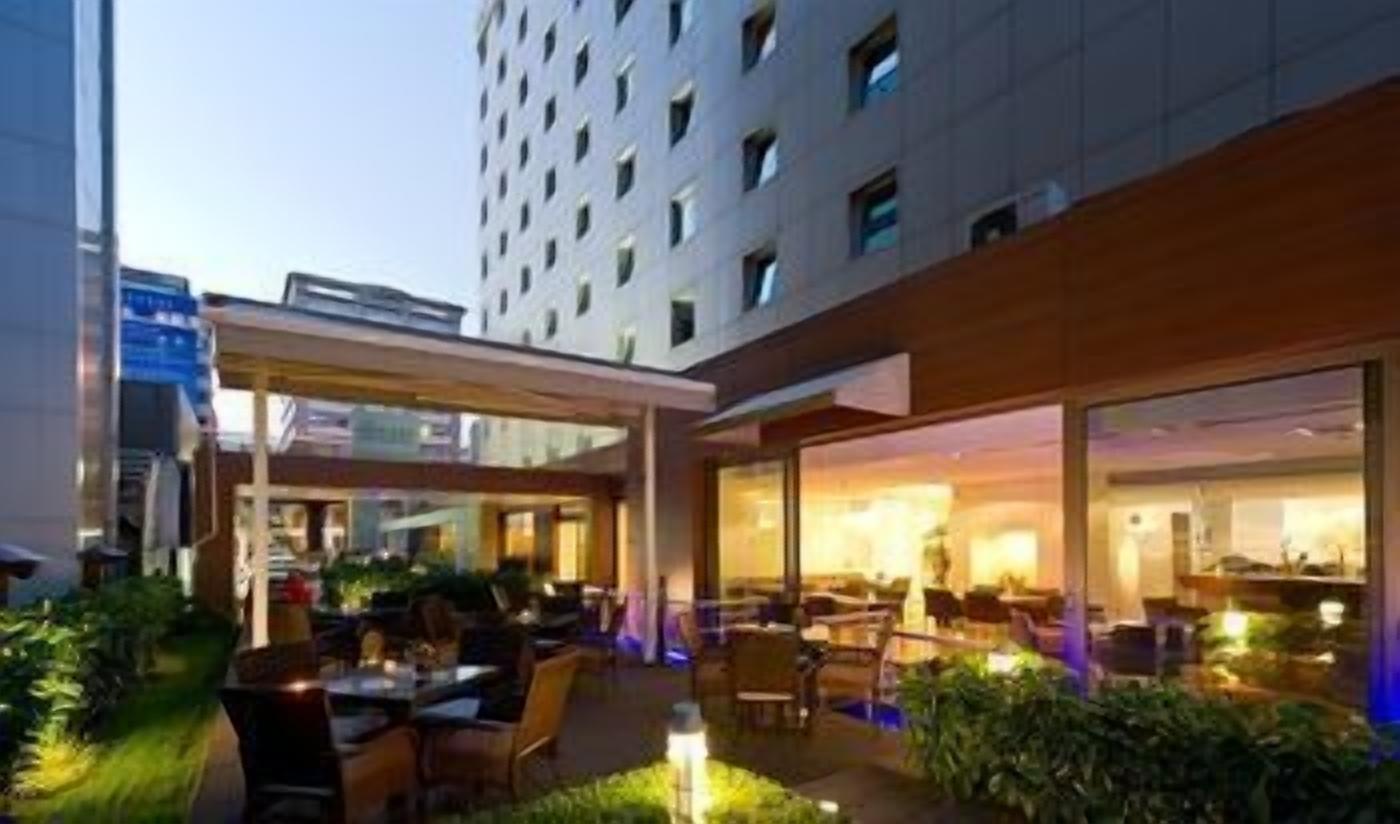 An outdoor dining area with tables and chairs, a building with multiple floors in the background, and a clear sky.