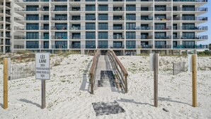 Un hotel de varios pisos con balcones, vista a la playa y un paseo marítimo de madera que conduce a la entrada.