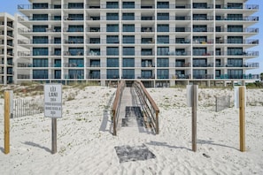Un hôtel à plusieurs étages doté de balcons, offrant une vue sur la plage, et une passerelle en bois menant à l’entrée.