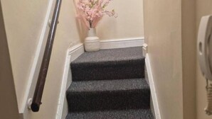 A staircase with carpeted steps, a white vase with pink flowers, and a handrail. - Glams one bedroom apartment (Wolverhampton)