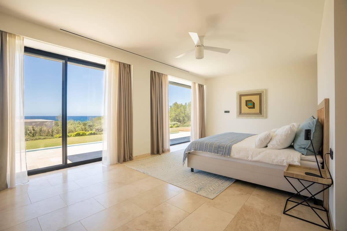 Ein Schlafzimmer mit Bett, Deckenventilator, Blick durch große Fenster und einem gerahmten Bild an der Wand.