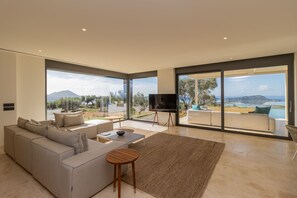 A modern living room with a large sofa, a coffee table, and a TV. There are large windows offering a view of the outdoors. - Messinian Nest (Pylos-Nestoras)